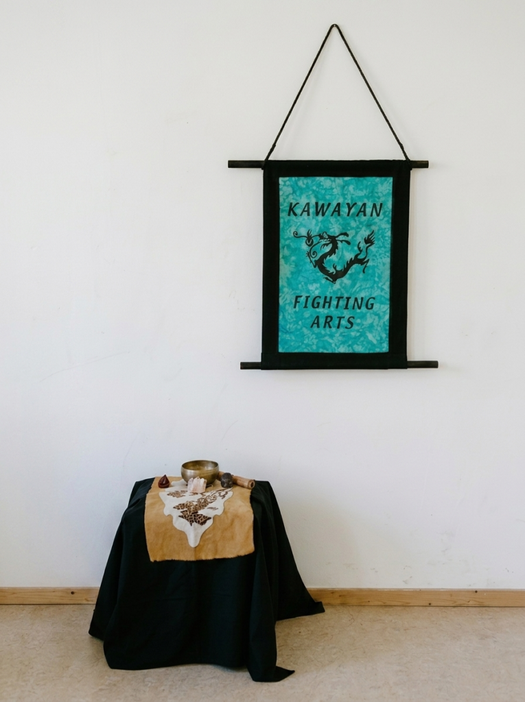 Kawayan Fighting Arts banner in teal and black with a dragon motif, hanging on a white wall above a draped table with a singing bowl, crystals, and a decorated hide cloth.